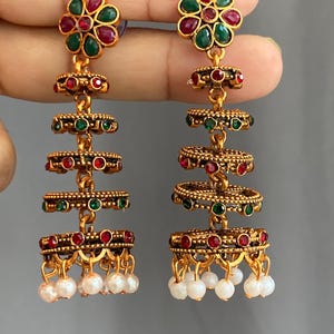 May include: A pair of gold-toned dangle earrings featuring a flower-shaped stud with red and green stones. The earrings have a tiered design with circular, textured metal discs accented by red and green stones, finished with dangling white beads.