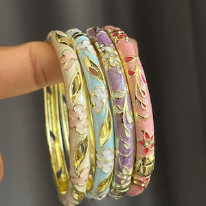 May include: A set of four hinged metal bangles featuring enamel floral patterns in cream, light blue, lavender, and pink. Each bracelet is accented with gold-tone metal vines and small clear stones embedded within the flower centres and stems.
