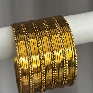 May include: A set of multiple gold-coloured metal bangles arranged in a stack. Each bangle features a smooth, reflective surface with a single thin band of textured, repeating geometric square patterns running along the centre of the circumference.