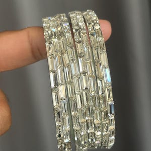 May include: A set of four silver-toned bangles, each featuring a continuous row of rectangular, clear, faceted stones set in a metallic frame. The bangles have a reflective, high-shine finish and are arranged side-by-side to display the stones.