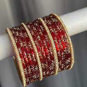 May include: A set of stacked bangles featuring deep red translucent glass rings accented with gold-toned metal edges and embedded clear crystals. The design includes alternating bands of plain red glass and red glass decorated with small, sparkling crystal floral patterns.
