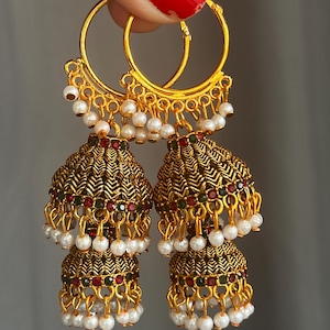 May include: A pair of gold-toned jhumka earrings featuring a double-tiered bell shape with intricate chevron metalwork. The design is accented with small red and green stones and finished with dangling white faux pearls along the bottom edges.