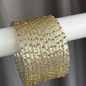 May include: A set of eight clear, translucent bangles featuring a textured, faceted surface. Each individual bangle is adorned with small, embedded metallic gold-toned studs arranged in a repeating pattern around the entire circumference.