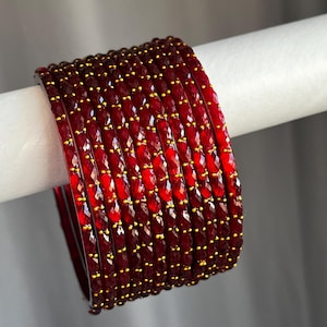May include: A set of ten deep red bangles featuring a faceted, textured surface with small, evenly spaced gold-coloured dots embedded along the outer rim of each individual bracelet, creating a uniform and reflective appearance across the stack.