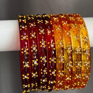 May include: A set of seven translucent bangles arranged in a gradient from deep red to bright yellow. Each bangle features a faceted geometric surface texture and is decorated with small, raised gold-tone dots and embedded clear crystals.