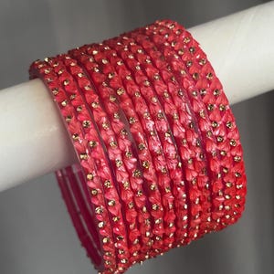 May include: A set of eight vibrant red bangles featuring a textured, petal-like surface pattern. Each bangle is adorned with small, evenly spaced gold-toned accents that catch the light, creating a consistent and repetitive decorative design.