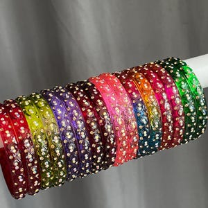May include: A collection of twelve circular bangles arranged in a row, each featuring a translucent coloured finish and a repeating embossed pattern embedded with small, reflective, iridescent crystals in shades of red, green, purple, and pink.