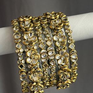 May include: A set of six gold-toned bangles featuring floral motifs and inset clear stones. Each bangle is decorated with alternating flower shapes and geometric patterns, with a metallic finish and reflective stone accents throughout the design.