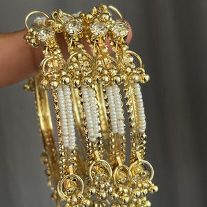 May include: A set of four gold-toned metal bangles featuring a central row of small white beads. Each bangle is accented with clear round crystals at the top and small dangling gold-toned metal spheres along the edges of the band.