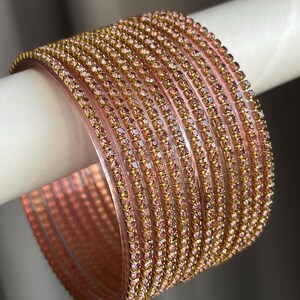 May include: A set of ten translucent pink bangles, each featuring a single row of small, sparkling pink rhinestones set in a gold-toned metal channel. The bracelets are stacked together, creating a uniform, shimmering appearance across the surface.