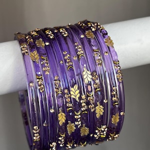 May include: A set of translucent purple bangles featuring gold-toned leaf motifs and small, sparkling embedded crystals. The circular bracelets are arranged in a stack, showcasing a smooth, glossy finish and consistent decorative patterns.