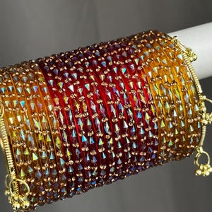 May include: A set of stacked bangles featuring a gradient of amber, orange, and deep red translucent glass. Each bangle is adorned with small, faceted, iridescent crystals and gold-toned metal accents, including small dangling bell charms.