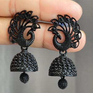 May include: A pair of matte black jhumka earrings featuring a peacock motif at the top with an intricate fan-shaped tail design. The bell-shaped base is textured with small circular patterns and finished with a small dangling beaded sphere.