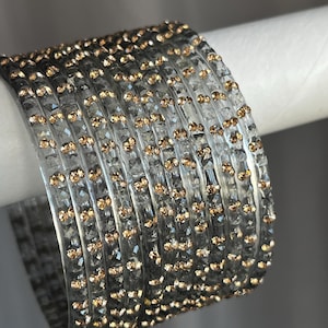 May include: A set of eight clear glass bangles featuring a faceted texture and embedded with small, round, amber-coloured crystals. The bracelets are arranged in a stack, showcasing the reflective quality of the glass and the warm sparkle of the stones.