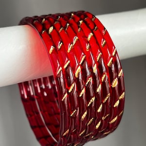 May include: A set of five translucent red bangles featuring a repeating diagonal gold-coloured pattern etched into the surface. Each circular bracelet has a smooth, rounded exterior and a consistent width, creating a stackable jewellery set.