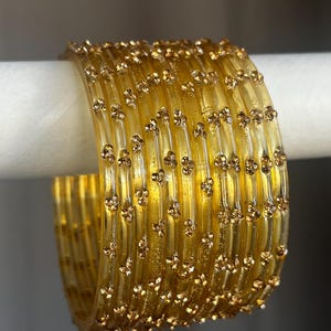 May include: A set of translucent yellow bangles featuring a repeating pattern of small, raised gold-toned floral accents. The circular bracelets are stacked together, showcasing a smooth, glossy finish and a consistent, uniform design across each piece.
