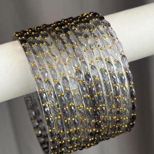 May include: A set of eight translucent grey bangles with a faceted texture and small, evenly spaced gold-coloured dots embedded along the surface. The bracelets are arranged in a stack, showcasing the repeating pattern and the clear, glass-like material.