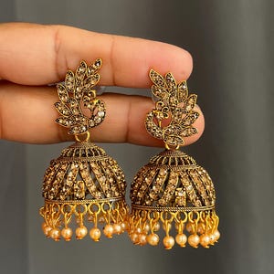 May include: A pair of gold-toned jhumka earrings featuring a peacock-shaped stud design encrusted with small clear crystals. The bell-shaped base is decorated with intricate metal filigree, embedded crystals, and a row of small dangling cream beads.