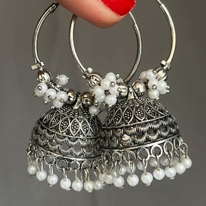 May include: A pair of silver-toned hoop earrings featuring bell-shaped jhumka pendants. The earrings are decorated with intricate metal filigree patterns and clusters of small, round white beads attached to the hoops and the bottom edges.