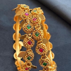 May include: Two gold-toned bangles featuring a series of circular, textured links. Each link contains a small, square-cut gemstone in alternating red and green colours. Small, flat, gold-toned leaf-shaped charms hang from the circular links.