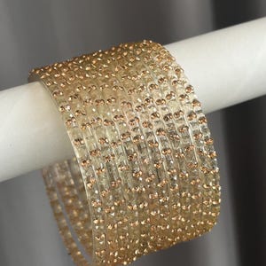 May include: A set of eight translucent, light-coloured bangles featuring a faceted surface texture. Each individual bangle is adorned with small, evenly spaced, metallic copper-toned accents embedded along the outer edge of the circular band.