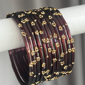 May include: A set of dark translucent purple bangles featuring a repeating pattern of small, sparkling gold-toned crystals arranged in floral-like clusters along the outer surface of each circular bracelet, creating a cohesive and decorative accessory.