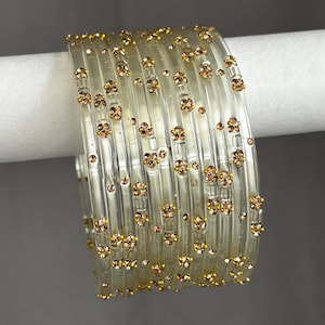May include: A set of clear, transparent bangles featuring small, scattered clusters of gold-toned rhinestones. The circular bracelets are arranged in a stack, showcasing a smooth, glossy finish and a repetitive floral-like pattern of crystals.