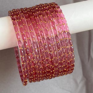 May include: A set of ten translucent pink bangles featuring a faceted surface texture. Each bangle is adorned with small, evenly spaced gold-coloured beads embedded along the outer edge, creating a repeating pattern of pink and gold accents.