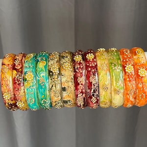 May include: A collection of twelve circular bangles arranged in a row. Each bangle features a unique marbled resin pattern in shades of yellow, red, turquoise, green, and orange, accented with small, gold-toned metal floral embellishments.
