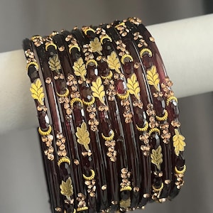 May include: A set of dark purple translucent bangles featuring gold-toned leaf motifs and small, sparkling light brown crystals embedded along the surface. The bracelets are arranged in a stack, showcasing a repeating pattern of metallic foliage.