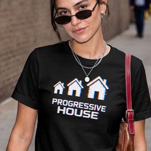 May include: A black short-sleeved t-shirt featuring a graphic of three white house outlines with a digital glitch effect and the text "PROGRESSIVE HOUSE" printed in white below the houses. The shirt is worn with a brown leather shoulder bag.
