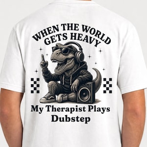 May include: A white t-shirt featuring a graphic of a Tyrannosaurus Rex wearing sunglasses, a hoodie, and headphones, sitting next to a speaker. The text reads: WHEN THE WORLD GETS HEAVY My Therapist Plays Dubstep. The design includes small star icons.