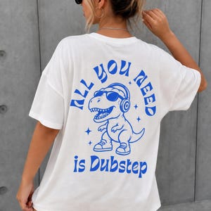 May include: A white short-sleeved t-shirt featuring a blue graphic on the back. The design depicts a cartoon dinosaur wearing sunglasses and headphones, surrounded by small star icons, with the text ALL YOU NEED is Dubstep in blue.