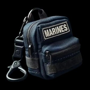 Può includere: Un portachiavi a forma di mini zaino in denim blu con cerniera nera e una toppa bianca con la scritta "MARINES". Il portachiavi ha un moschettone nero e un anello metallico per attaccarlo a chiavi o borse.