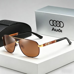 Op de afbeelding: Bruine piloten zonnebril met gouden montuur en zwarte pootjes. De zonnebril heeft een bruine tint en wordt gepresenteerd met een zwart etui en een zilveren doos met het Audi-logo.