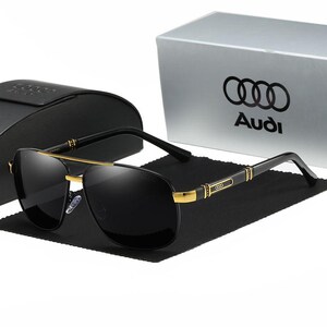 Op de afbeelding: Zwarte aviator zonnebril met gouden accenten en een zwart montuur, op een zwarte doek. De zonnebril ligt naast een zwarte etui en een zilveren doos met het Audi-logo en tekst.