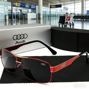 May include: Red and gold-framed sunglasses with black lenses. The sunglasses have black temples and the Audi logo on the temple and lens. The sunglasses are displayed on a black cloth, with a silver box and a black box in the background.