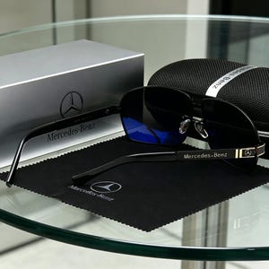 May include: Black sunglasses with blue lenses, branded with the Mercedes-Benz logo on the arms and temples. The glasses are displayed on a black cloth with the Mercedes-Benz logo, alongside a silver box and a black case.