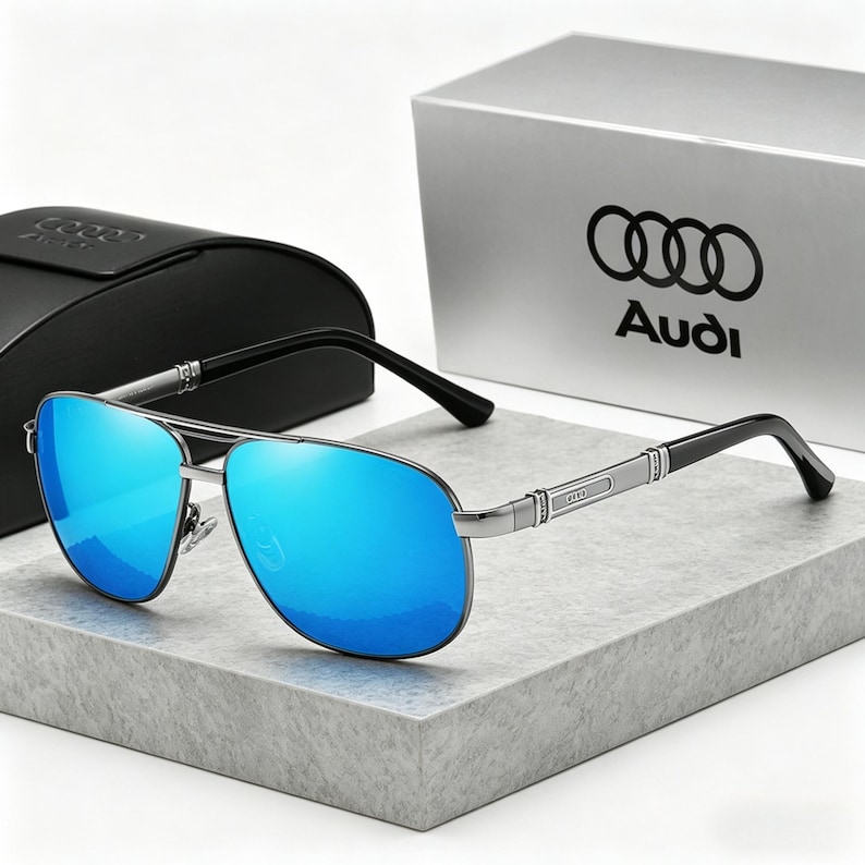 Op de afbeelding: Zilverkleurige piloten zonnebril met blauw gespiegelde glazen. De zonnebril heeft zwarte veren en een zilveren brug. De etui en doos op de achtergrond hebben het Audi-logo.