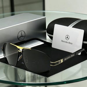 May include: Black sunglasses with gold detailing and the Mercedes-Benz logo. The sunglasses are presented with a case, cleaning cloth, and a card bearing the Mercedes-Benz logo. The lenses are dark, and the frames are black and gold.