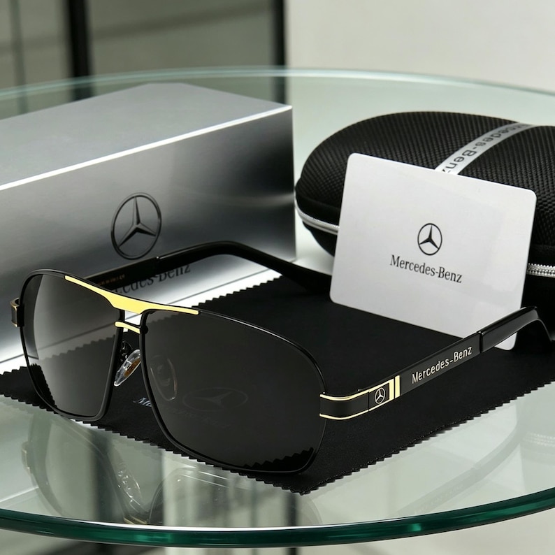 May include: Black sunglasses with gold detailing and the Mercedes-Benz logo. The sunglasses are presented with a case, cleaning cloth, and a card bearing the Mercedes-Benz logo. The lenses are dark, and the frames are black and gold.