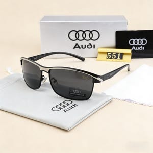 May include: Black and silver sunglasses with the Audi logo on the frame and lenses. The sunglasses are displayed with a white cleaning cloth, a gray pouch with the Audi logo, and a silver box with the Audi logo.