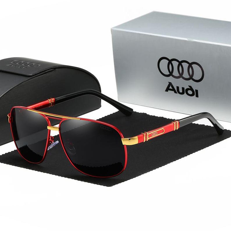 Op de afbeelding: Rode en gouden piloten zonnebril met zwarte glazen. De zonnebril heeft een rood montuur met gouden accenten en zwarte veren. Een zwarte etui en een zilveren doos met het Audi-logo staan op de achtergrond.