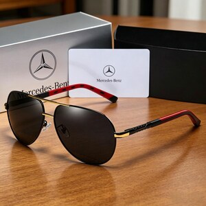 May include: Black aviator sunglasses with gold frames and red accents on the temples. The sunglasses are displayed with a box and card, both featuring the Mercedes-Benz logo and text.