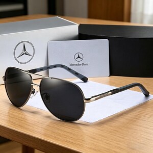 May include: Aviator-style sunglasses with dark lenses and a silver frame, resting on a white cloth. The sunglasses are next to a card and box with the Mercedes-Benz logo.