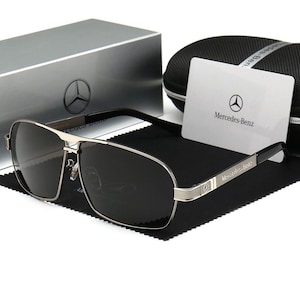 May include: Silver-framed sunglasses with dark lenses, featuring the Mercedes-Benz logo on the temple and a matching case. The sunglasses rest on a black cloth, with a card bearing the brand's emblem.