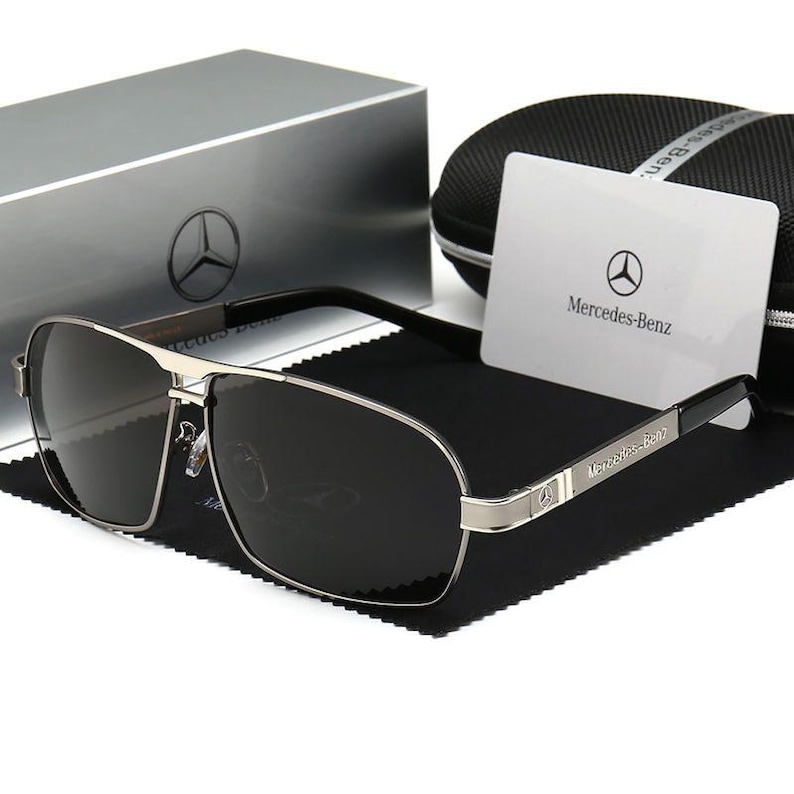 May include: Silver-framed sunglasses with dark lenses, featuring the Mercedes-Benz logo on the temple and a matching case. The sunglasses rest on a black cloth, with a card bearing the brand's emblem.