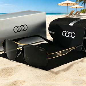 May include: Black sunglasses with gold accents and the Audi logo on the frame and case. The sunglasses are displayed on a sandy beach with a case and box in the background.