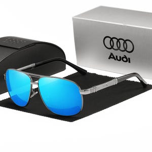 Op de afbeelding: Zilverkleurige zonnebril met blauwe spiegelglazen. De zonnebril heeft een klassieke pilotenstijl met zwarte pootjes. Een zwarte etui en een witte doos met het Audi-logo staan op de achtergrond.