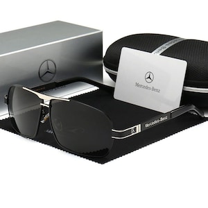 May include: Black sunglasses with a silver frame and dark lenses. The sunglasses are displayed with a case, cleaning cloth, and a card that says "Mercedes-Benz". The sunglasses have the Mercedes-Benz logo on the frame.